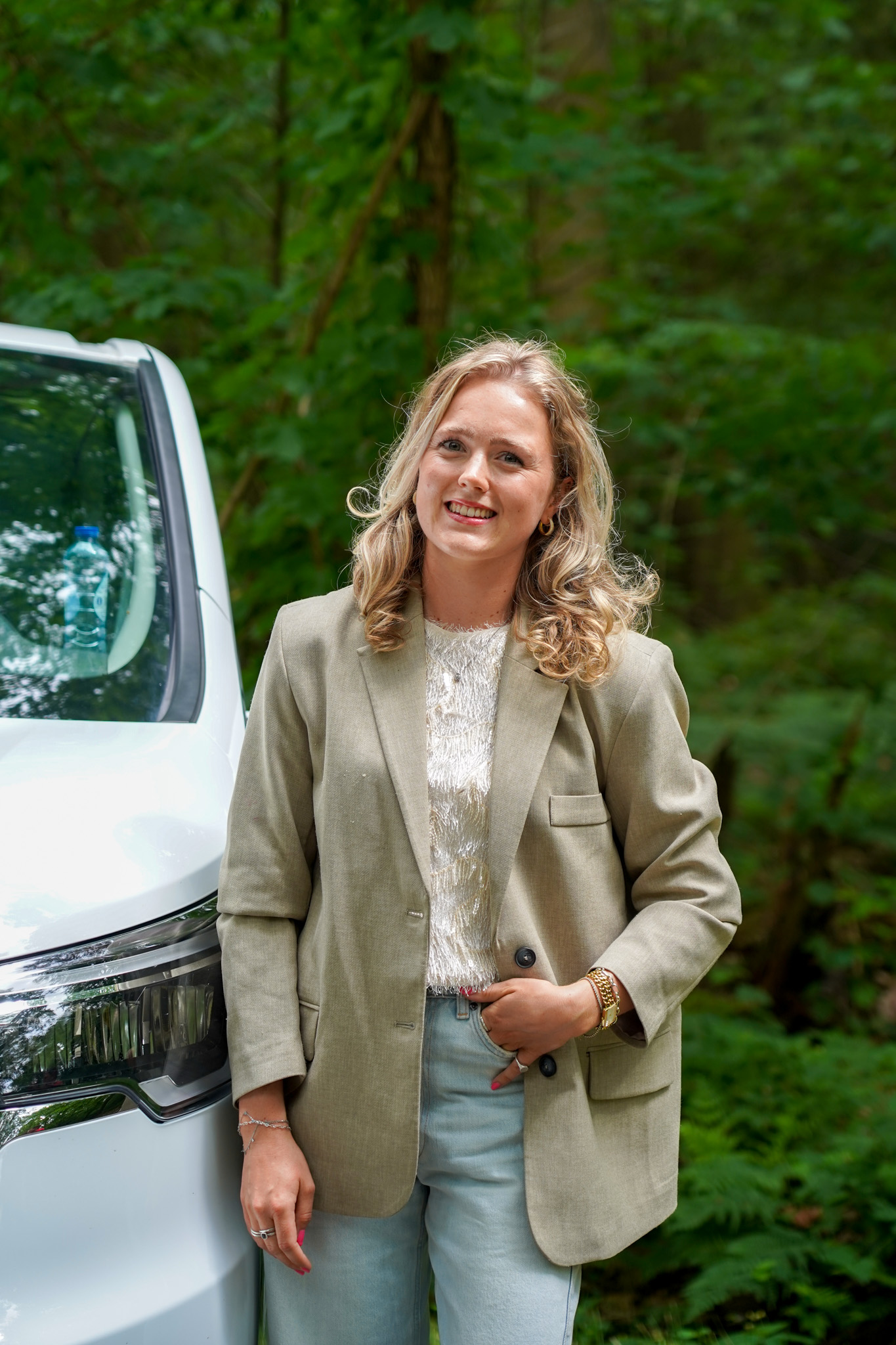 Vrouw in beige blazer en lichtblauwe jeans staat naast witte bedrijfsauto voor groen geboomte.