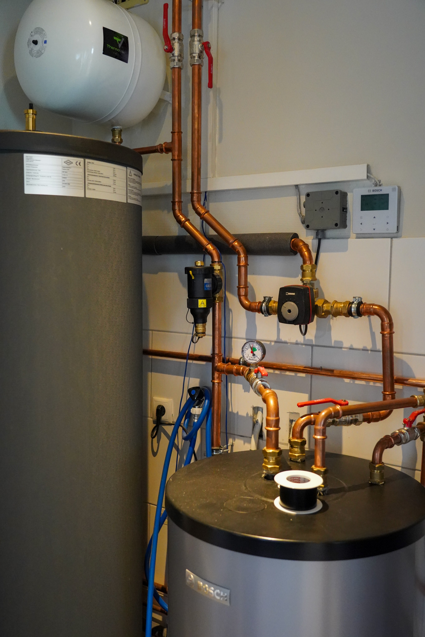 Moderne verwarmingsinstallatie met Bosch boiler, expansievat, circulatiepompen en thermostaat tegen witte wand met koperen leidingen.