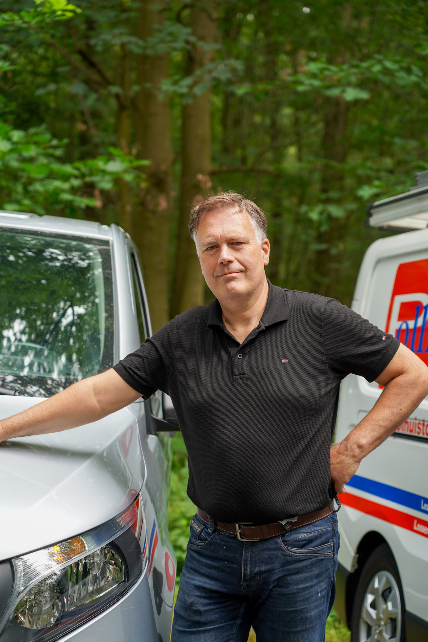 Man in zwart poloshirt leunt tegen witte bestelwagen voor groene bomen.