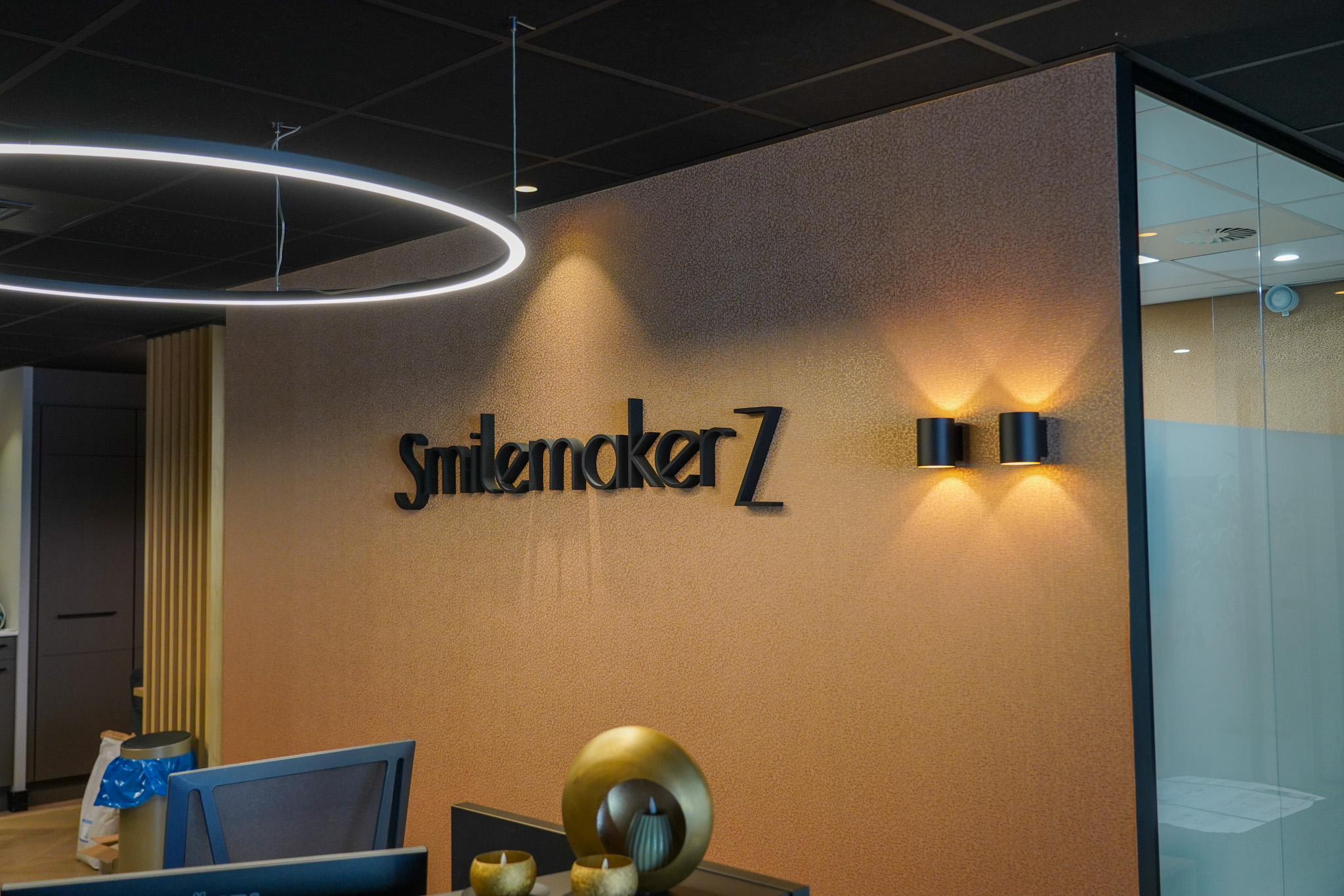 SmilemakeZ logo aan gouden muur met neon ring verlicht, moderne kantoorinrichting met glas en design elementen.