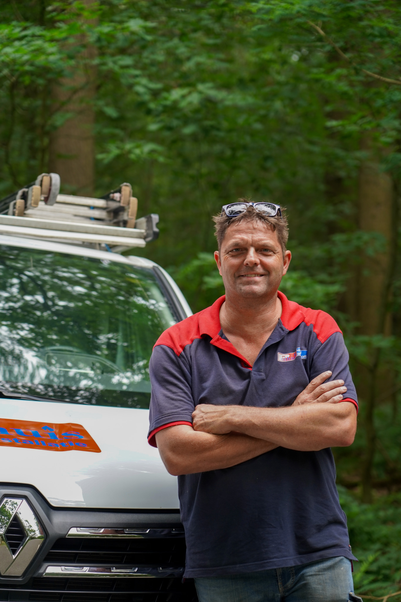 Installateur van Polhuis staat met gekruiste armen voor bestelwagen met dakrek in bosrijke omgeving.